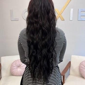 A woman with long wavy hair styled by Loxxie, dressed in a red plaid shirt, looking cheerful.