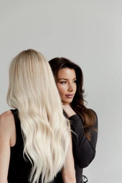 Two women are standing close together against a gray background. One has long, straight blond hair, while the other has dark wavy hair, creating a contrasting visual.