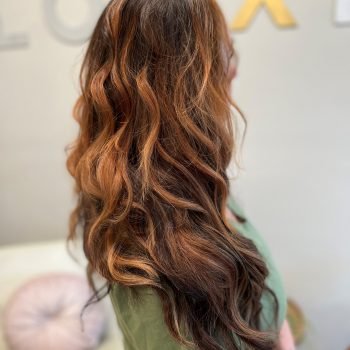 A woman with long wavy Loxxie hair, wearing a red plaid shirt, stands against a neutral background.
