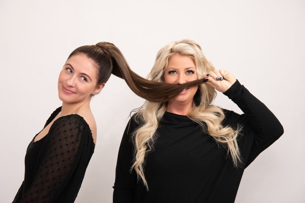 Two women in black shirts are playfully posing against a white background. One holds her long, dark ponytail up; the other smiles with it against her face.