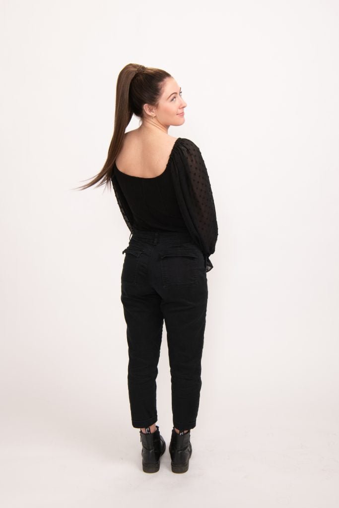A woman with a ponytail, wearing a black outfit, stands with her back to the camera in a minimal white space, conveying a calm and introspective mood.