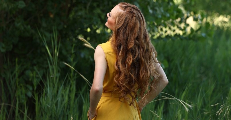 A woman with long red hair stands in a field, showcasing Loxxie Hair products against a natural backdrop.