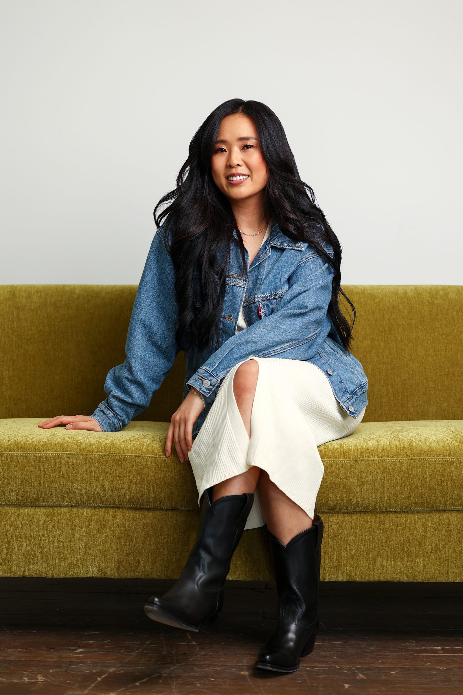 A woman in a denim jacket and white dress sits on a green couch, showcasing Loxxie Hair.