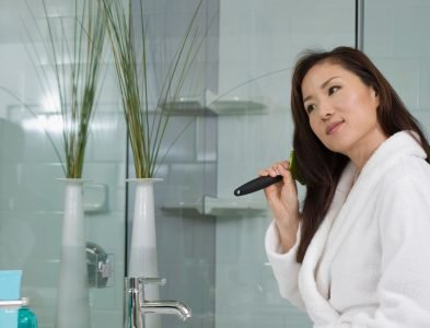 A woman is brushing her teeth in front of a mirror, showcasing her Loxxie Hair while maintaining her oral care.