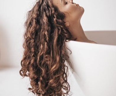 A woman with long curly hair relaxes in a bathtub, showcasing her Loxxie Hair products.